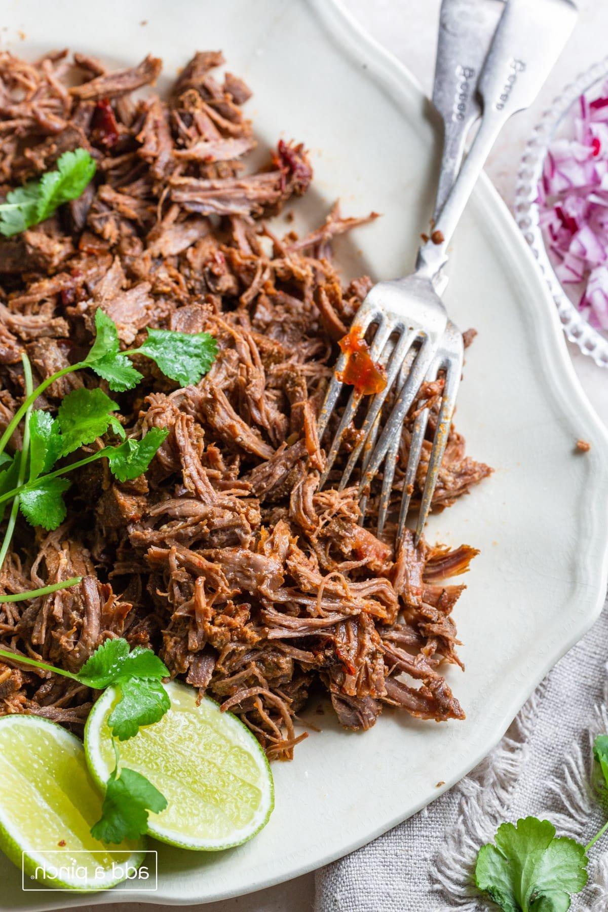 Barbacoa σε πιατέλα με κόλιανδρο, φέτες λάιμ και δύο πιρούνια.