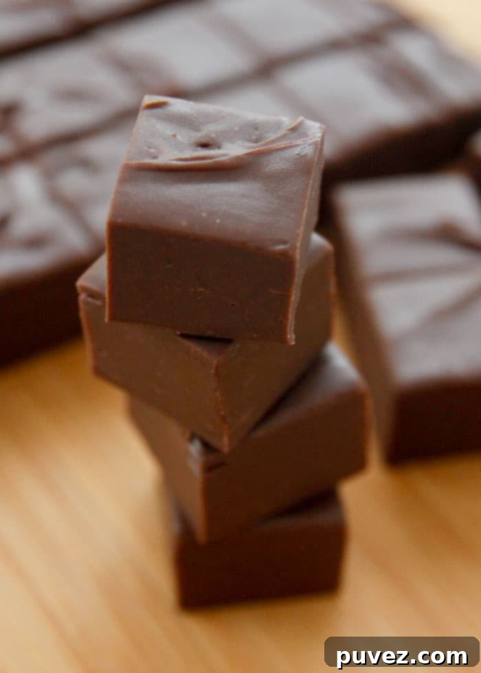 Four bite-sized pieces of chocolate fudge stacked on a wooden board with a slab of fudge in the background, showcasing its rich, dark color and smooth texture.