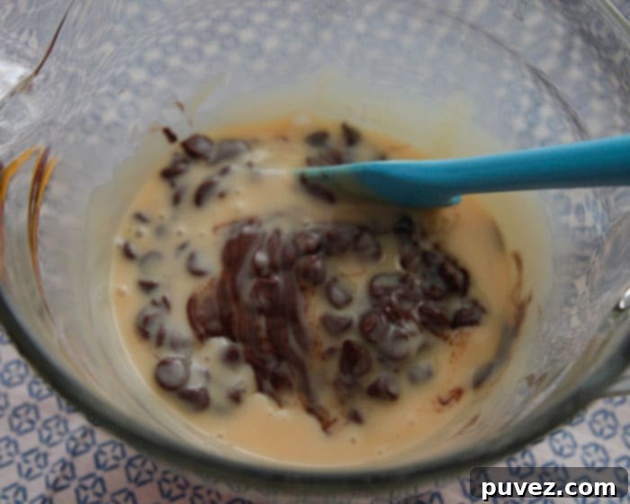 A vibrant blue spatula stirs sweetened condensed milk and partially melted chocolate chips in a clear glass bowl, showing the initial stages of the 3-minute fudge preparation.