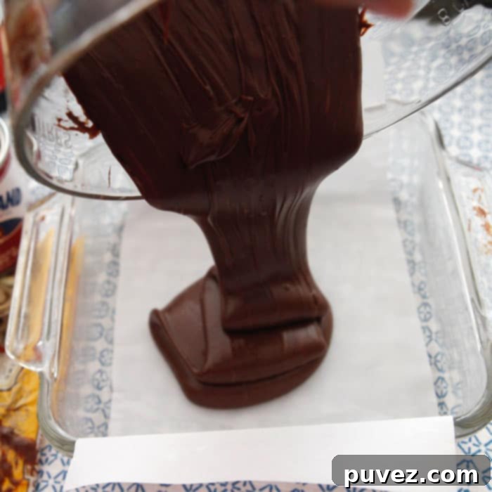 Three minute fudge mixture is poured from the glass bowl into a parchment paper lined square glass baking pan, showing the smooth, dark chocolate flowing easily.