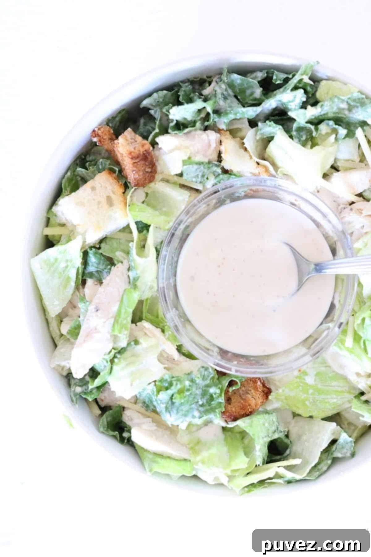 A small glass bowl with Caesar dressing with a spoon in the middle of a bowl of Caesar salad.