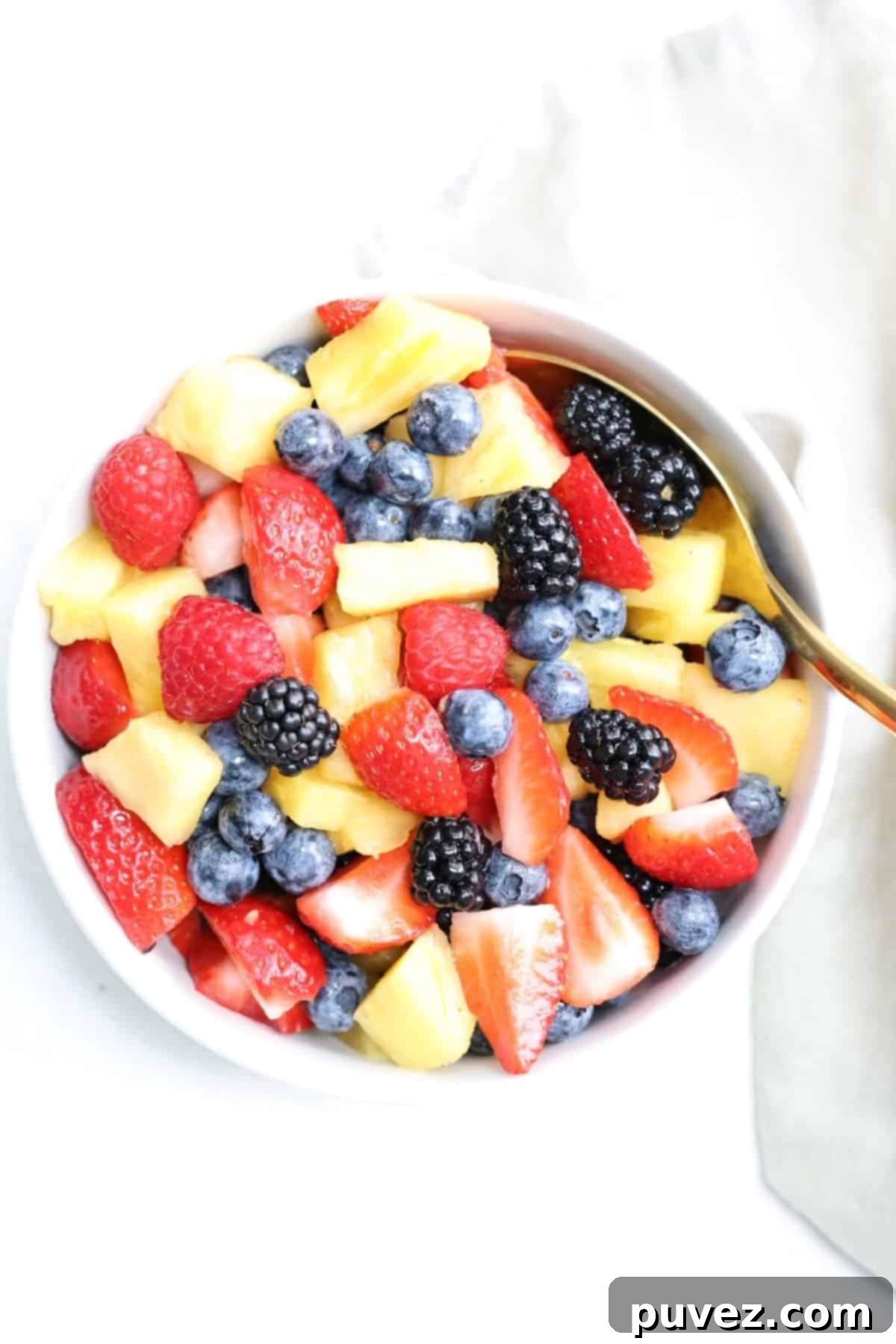 Top view of a bowl of fresh fruit salad with a gold spoon and linen napkin to the side.