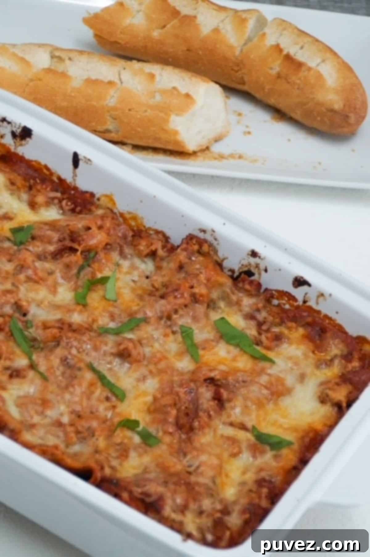 Baked lasagna with two halves of a baguette on a white plate behind the lasagna.