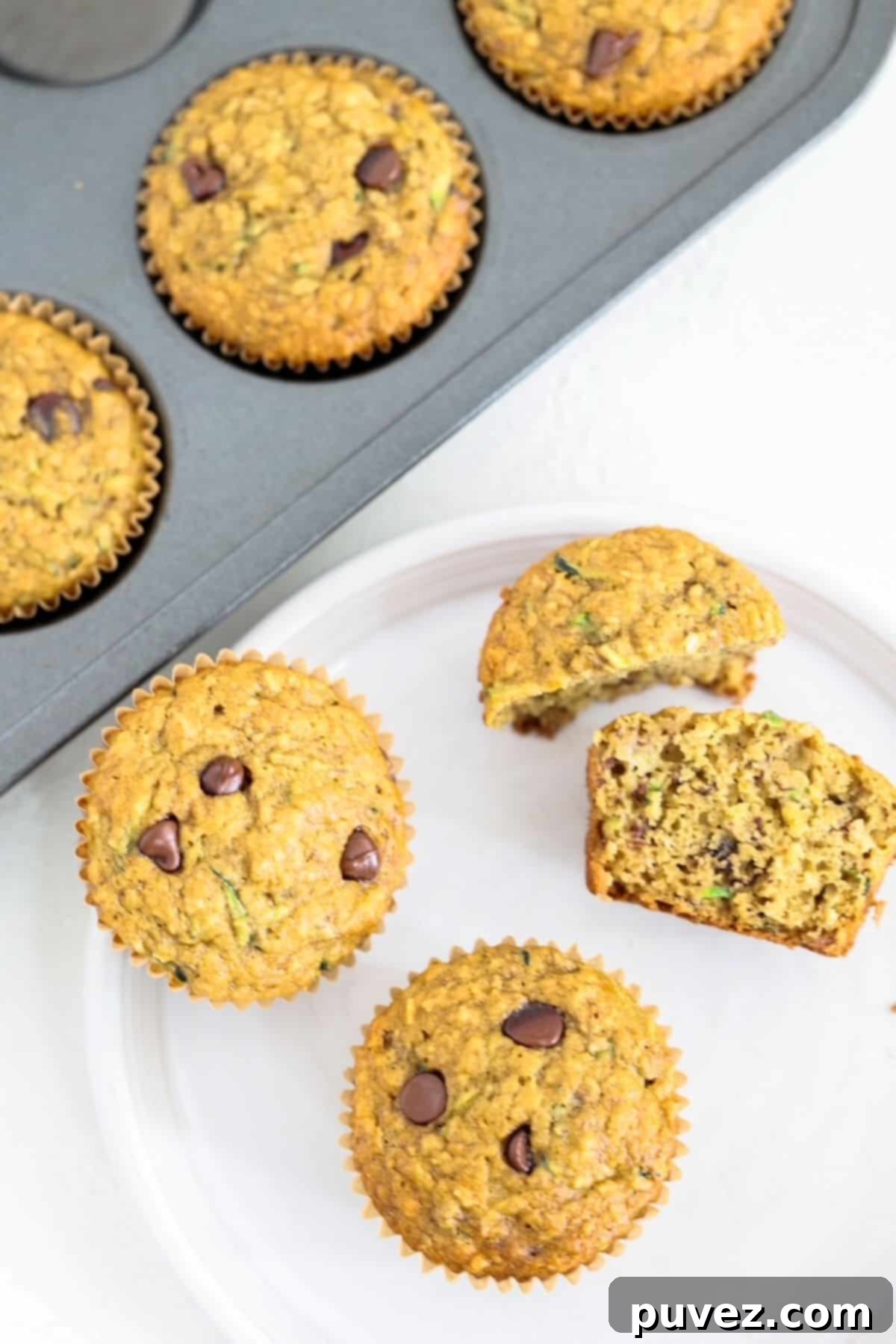 Two whole and two halves of banana chocolate chip muffins on a whit plate with a muffin tin of muffins behind it.