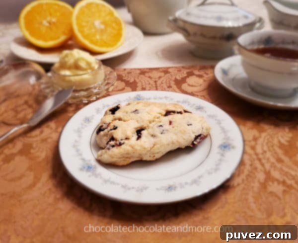 A place setting of fine china featuring a perfectly baked cranberry scone, generously topped with melted orange butter, alongside a steaming cup of tea and a fresh orange slice.