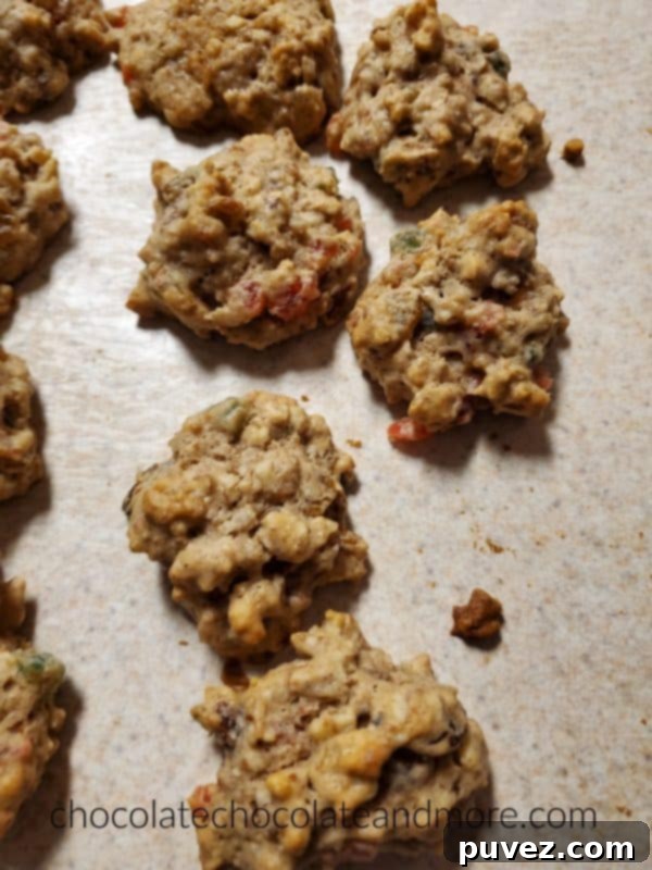 A close-up of several homemade fruitcake cookies, showing their moist texture and colorful bits of red and green candied fruit. The cookies are scattered on a kitchen counter, ready to be enjoyed or shared.