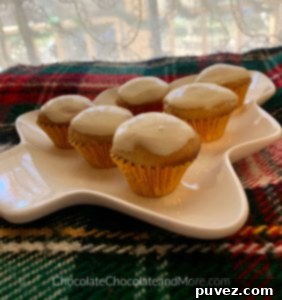 Six mini Eggnog Cream Cheese Cupcakes arranged artfully on a festive green, red, and white plaid blanket. A soft white lace curtain provides a charming, cozy background, highlighting the delicate frosting and sprinkle of spice on each cupcake.