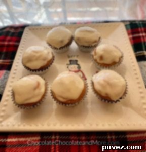 Seven regular-sized Eggnog Cream Cheese Cupcakes beautifully displayed on a square plate adorned with a snowman design. The plate rests on a vibrant red, green, and white plaid blanket, with a delicate white lace curtain softening the background, creating a charming winter scene.