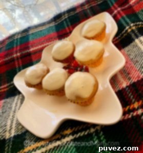 Six mini Eggnog Cream Cheese Cupcakes are artfully arranged on a cozy green, red, and white plaid blanket. A soft, white lace curtain hangs gently in the background, creating a warm and inviting atmosphere that perfectly complements these festive treats.