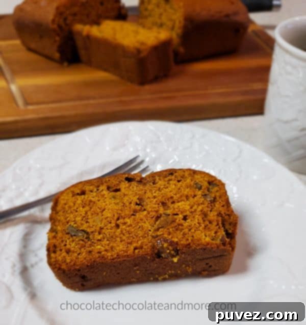 A perfectly baked slice of moist Pumpkin Raisin Bread on a white plate, with a full loaf in the background, showcasing its delicious texture and golden raisins. Perfect for a cozy breakfast.