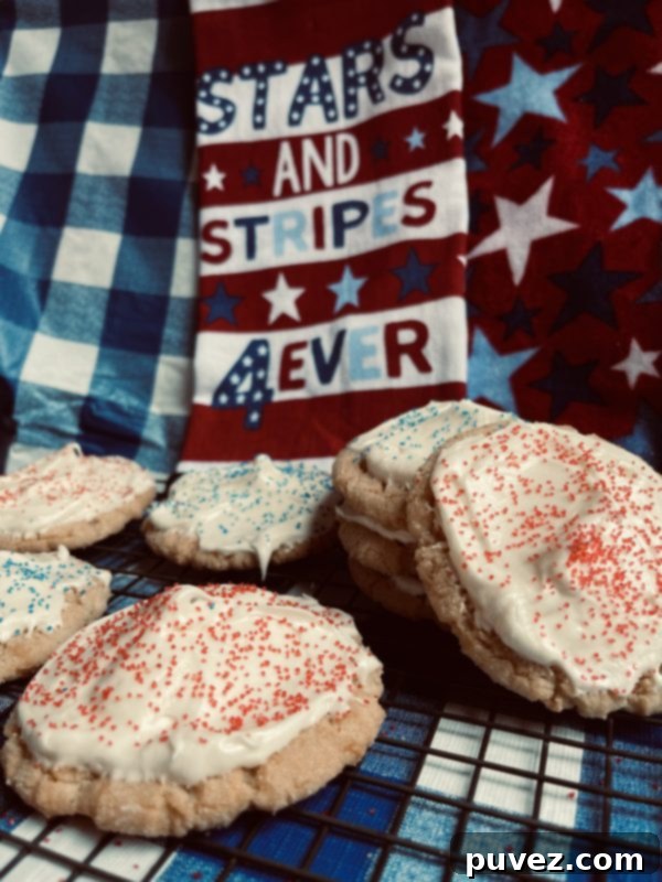 8 Cream Cheese Frosted Sugar Cookies sitting on a cooling rack on a checkered blue and white background with a "stars and stripes for ever" towel in the back.
