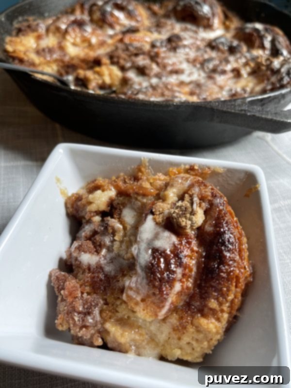 A serving of the Cinnamon Roll Dessert Casserole in a small dish, with the full skillet of the baked casserole in the background, showcasing its gooey layers and golden-brown crust.
