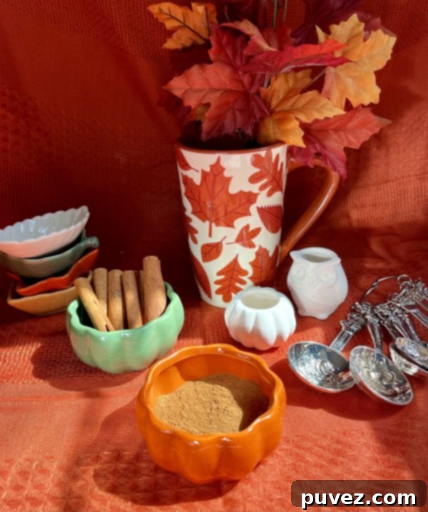 All the freshly mixed pumpkin pie spice ingredients are now combined in an inviting orange pumpkin-shaped bowl. In the background, leaf-shaped bowls are neatly stacked, and a larger pumpkin bowl holds cinnamon sticks. A white ceramic pumpkin and an owl figurine, alongside measuring spoons and a festive fall mug with leaves, complete the autumn scene.