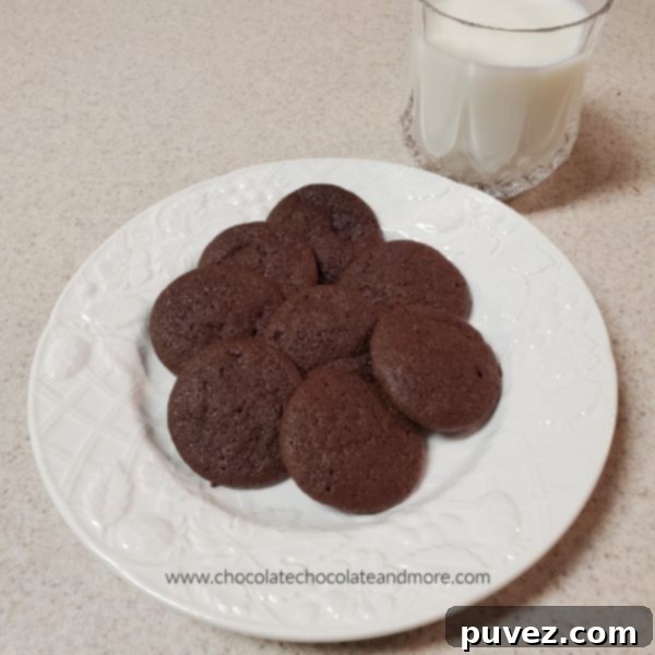 Several dark chocolate cookies presented beautifully on a white plate, positioned next to a refreshing glass of milk, highlighting their perfect dippability.