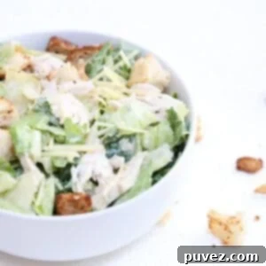 One chicken Caesar salad in a white bowl with croutons to the right of the bowl.