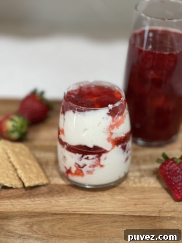 Beautifully layered Strawberry Shortcake Dip in a cup, served alongside graham crackers, fresh strawberries, and a taller cup of strawberry syrup on a wooden cutting board.