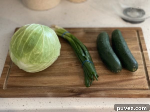Fresh, vibrant ingredients for the Green Goddess Salad laid out on a clean white surface, showcasing a head of cabbage, cucumbers, and green onions ready for chopping.