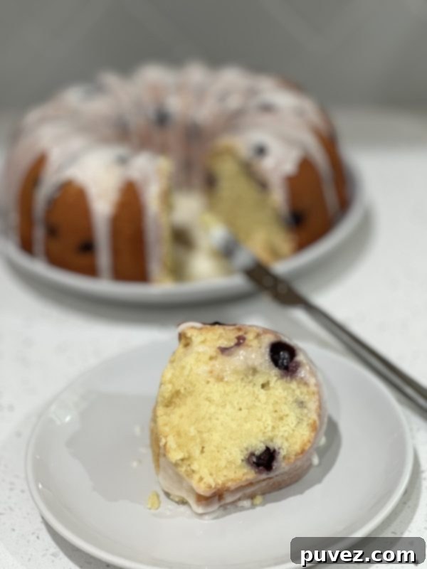 A beautiful slice of Lemon Blueberry Pound Cake on a white plate, showcasing its moist texture and blueberry specks, topped with a luscious lemon glaze.