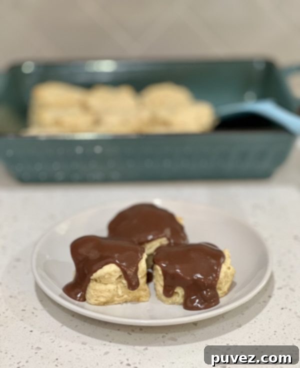 3 Chocolate Gravy Covered Buttermilk Biscuits on a white plate with the baking dish of biscuits behind it, highlighting the fluffy texture and rich chocolate