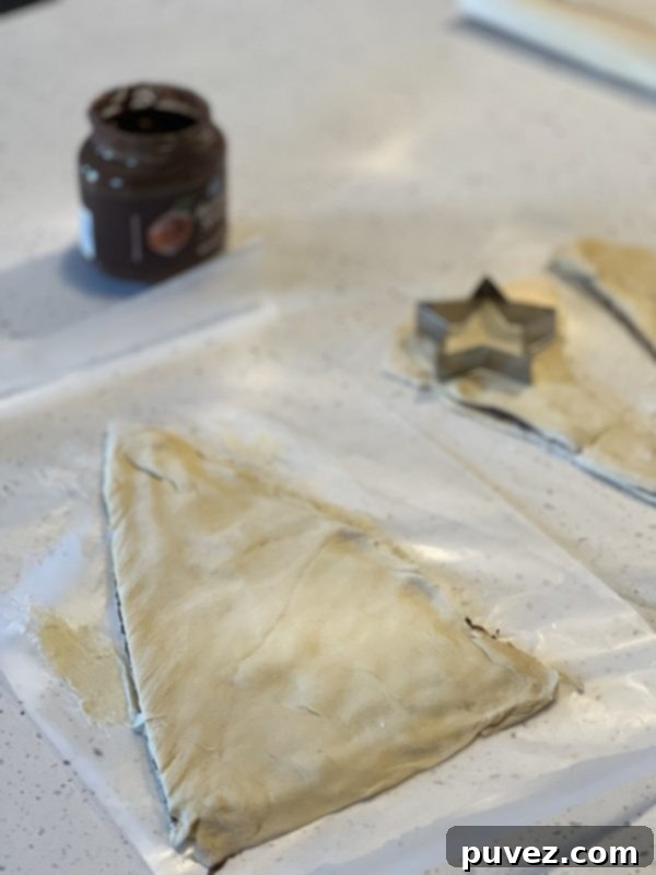 Layered crescent dough being cut into a Christmas tree shape with a knife, and a star cookie cutter pressing into the dough scraps to create the tree topper.