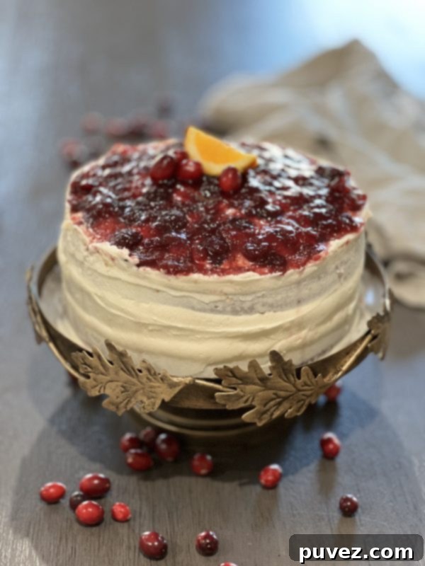 A full Cranberry Orange Triple Layer Cake on an elegant cake stand, beautifully garnished with fresh cranberries and orange slices, set on a wooden table.