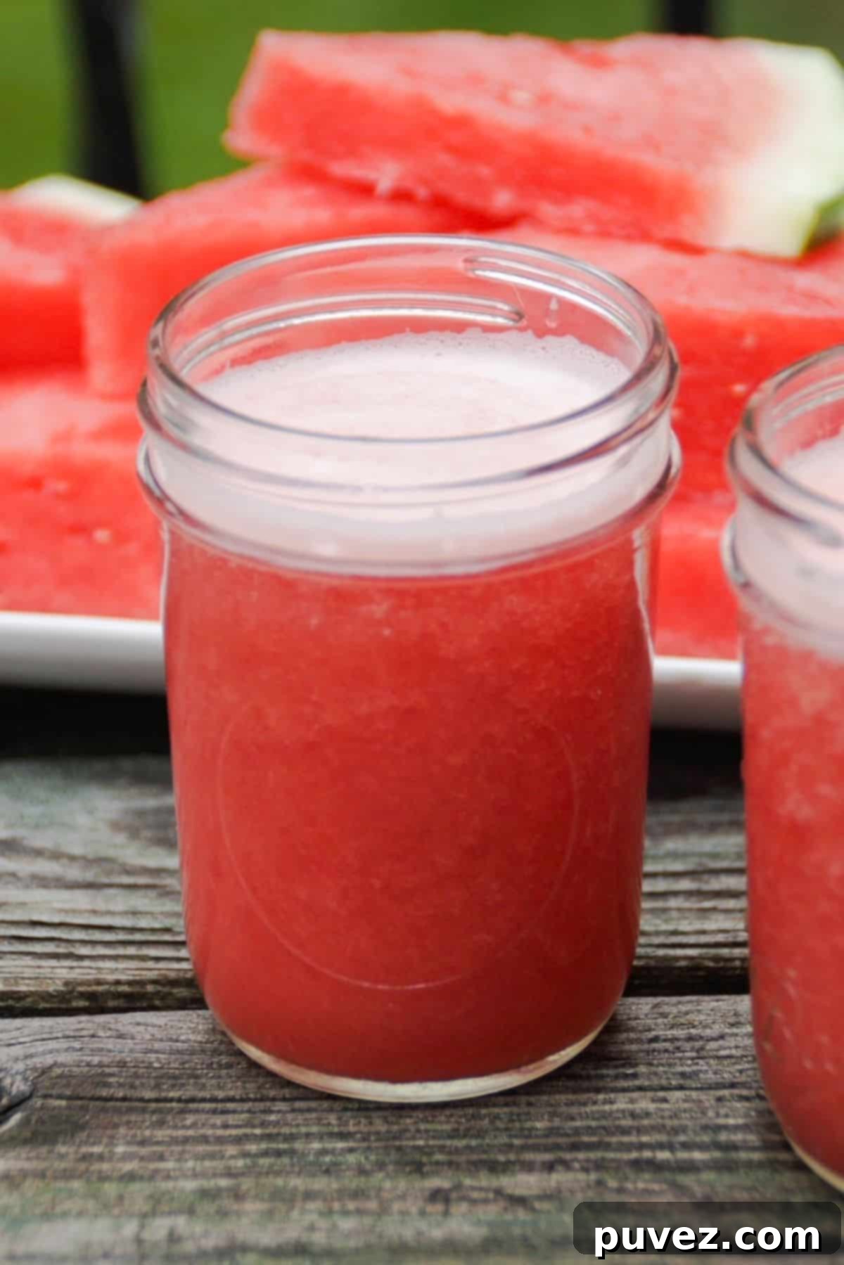 One watermelon banana smoothie in a small mason jar in the center with the side of a second smoothie to the right. A plate of stacked watermelon slices is behind the smoothies.