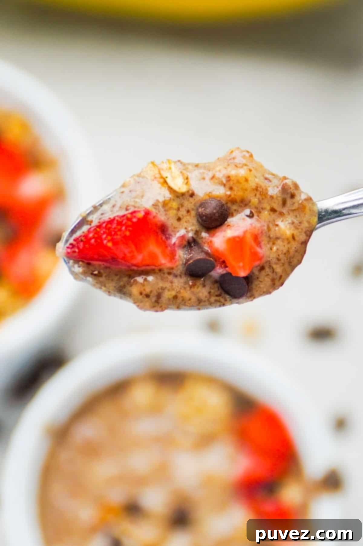 A close-up of a spoonful of creamy peanut butter banana flaxseed pudding, generously topped with small strawberry pieces and mini chocolate chips, ready to be enjoyed. In the soft background, two more small bowls of the same delicious pudding await, hinting at a shared, healthy treat.