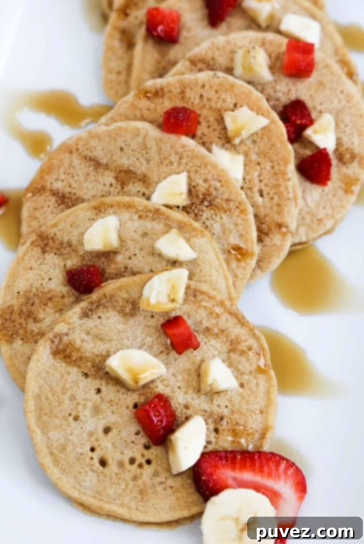 Several small pancakes overlapping, topped with pieces of banana and strawberry. Drizzled with maple syrup.