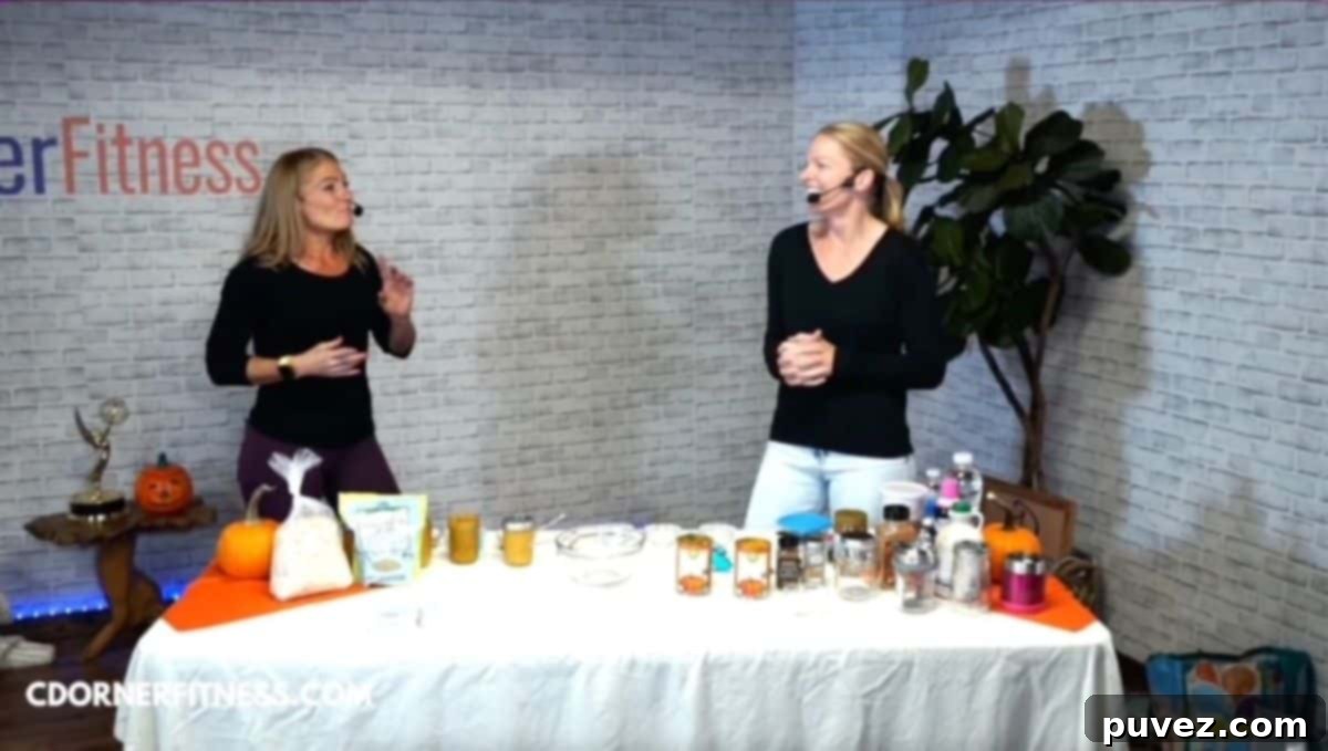 Chris Dorner and Kristi Ruth talking to each other while standing behind a table with a white tablecloth that has pumkin overnight oat ingredients as well as cooking utensils and food storage containers set up for a cooking demo. CDorner Fitness logo is on the back wall to the left. 