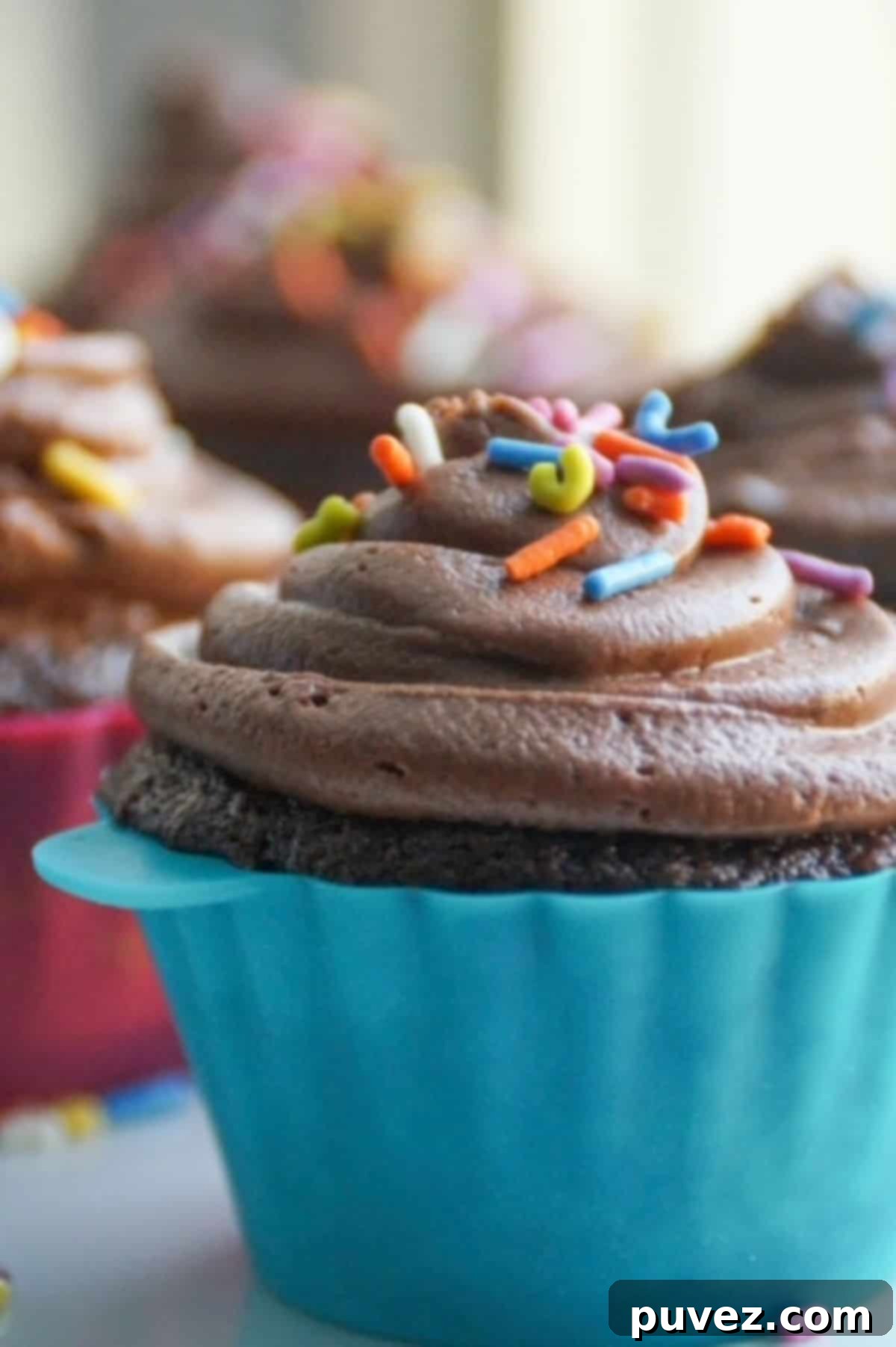 Chocolate frosted chocolate cupcake in a turquoise silicone cupcake liner and topped with sprinkles. More cupcakes are in the distant background. 