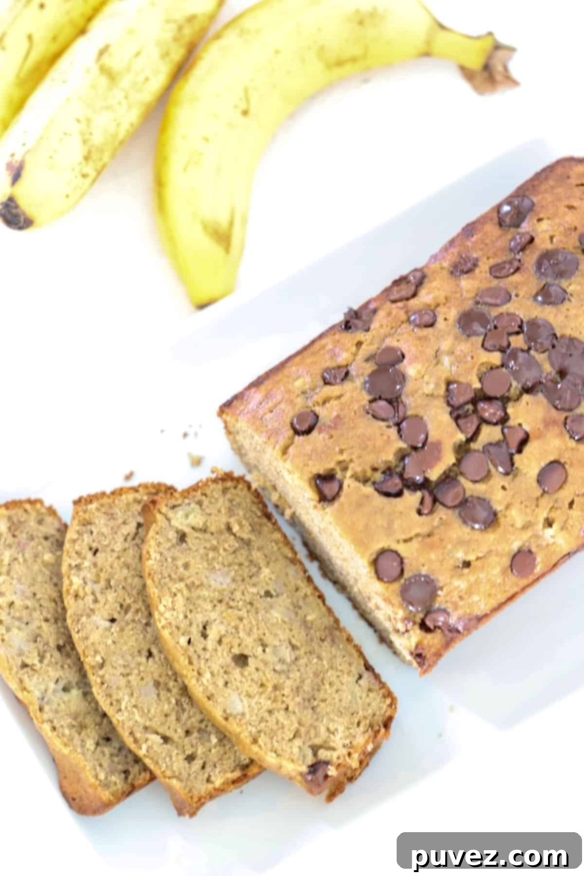 A loaf of banana bread partially sliced on a white platter with bananas in the background.