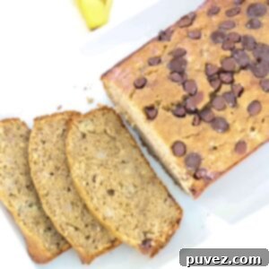A sliced loaf of dairy-free banana bread on a white platter, with ripe bananas in the background.