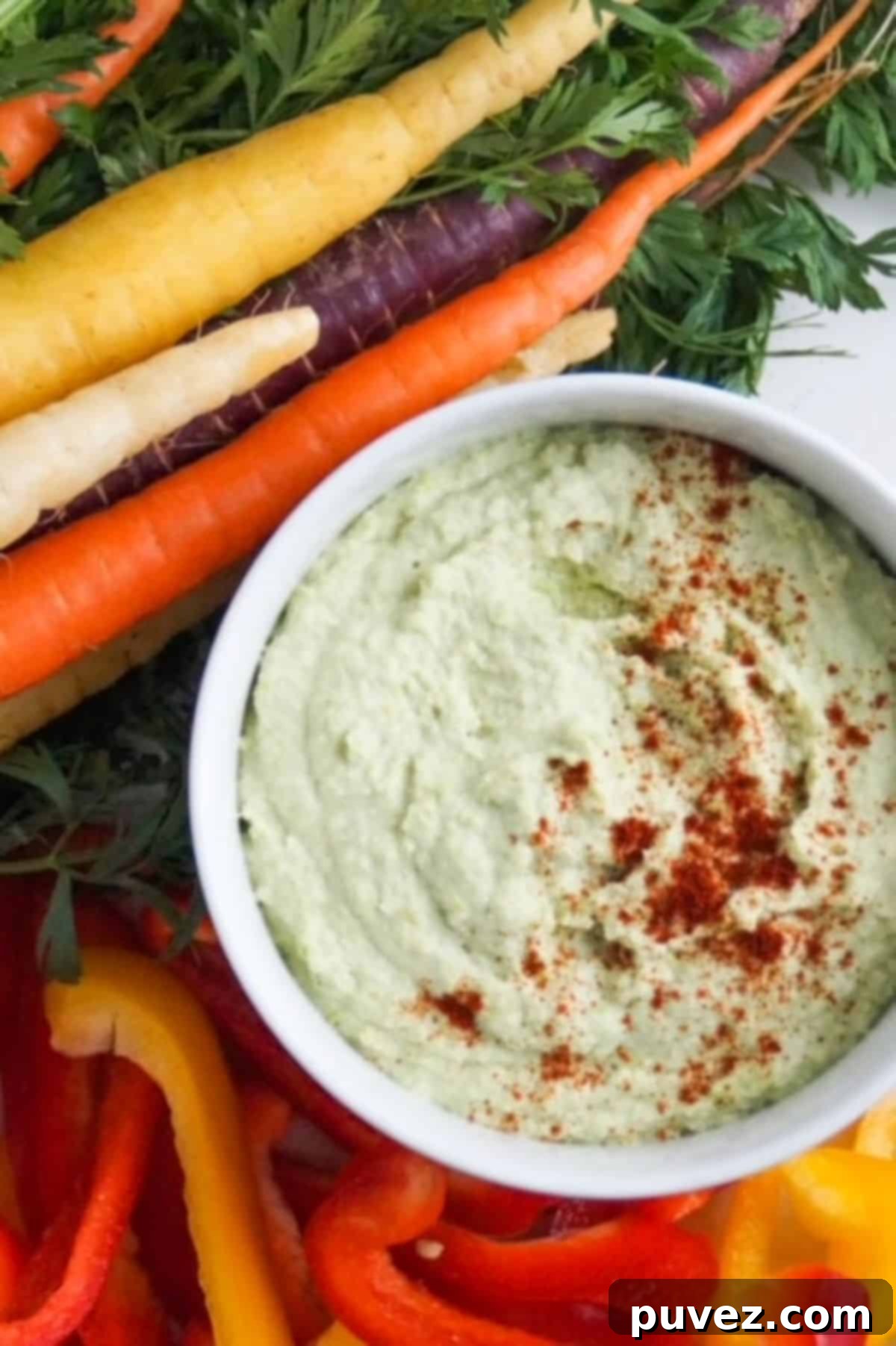 Edamame hummus in a white bowl, garnished with paprika, surrounded by tri-colored carrots and sliced red and yellow bell peppers. A perfect healthy snack.