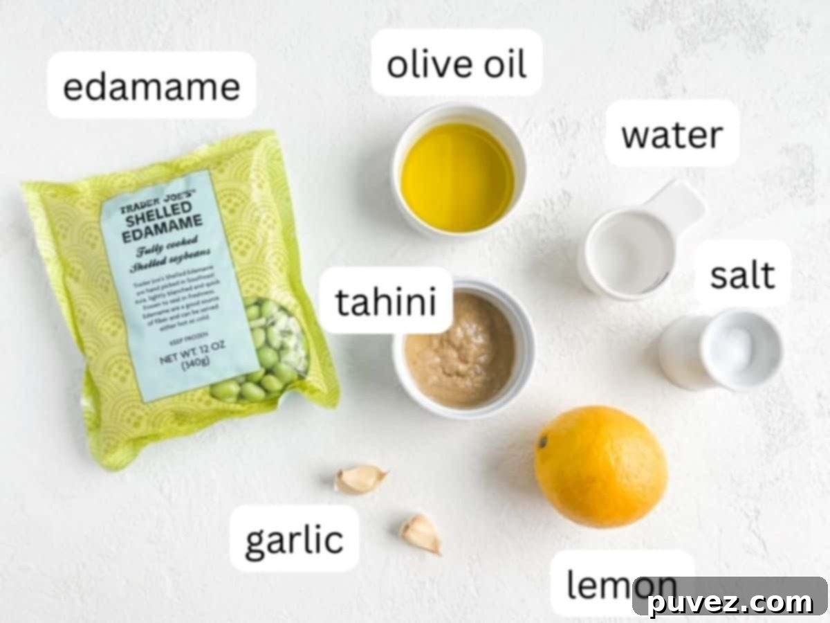 Individual ingredients for edamame hummus: a bag of shelled edamame, a whole lemon, two cloves of garlic, and small dishes of olive oil, water, tahini, and salt.