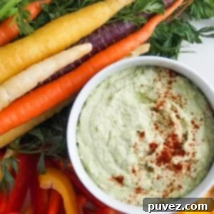 Edamame hummus in a white serving bowl, topped with paprika, surrounded by vibrant tri-colored carrots and slices of fresh bell pepper.