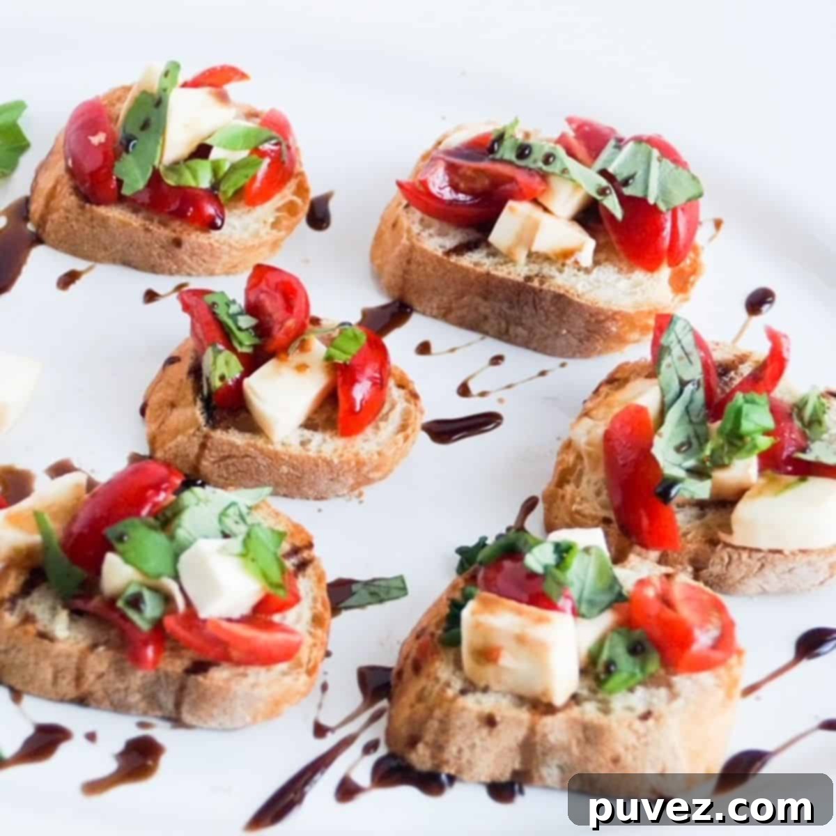 Six pieces of caprese bruschetta drizzled with balsamic glaze on a white platter. 