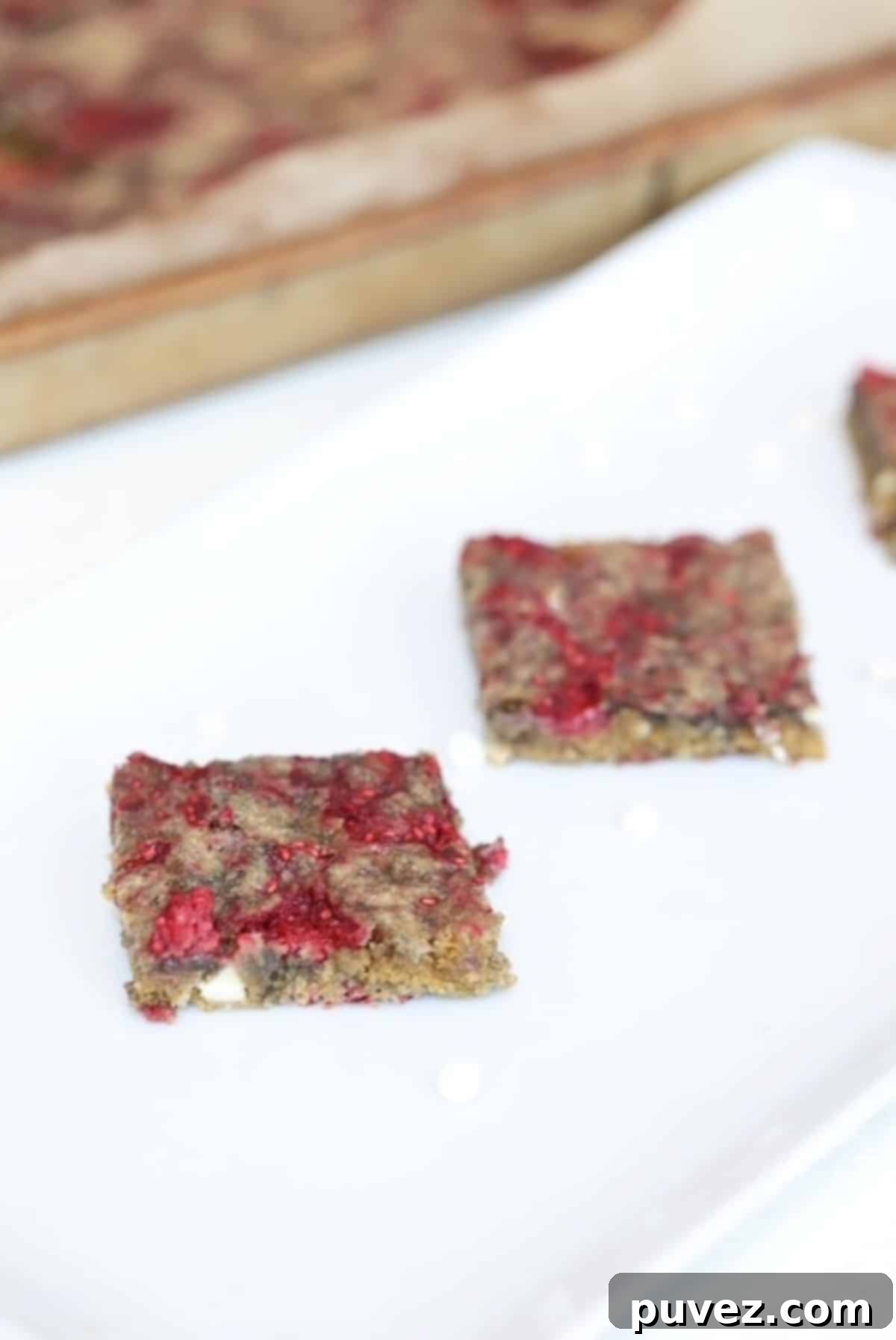 Two perfectly cut raspberry white chocolate blondies on a elegant white serving platter, with a baking pan filled with more blondies in the soft-focus background.