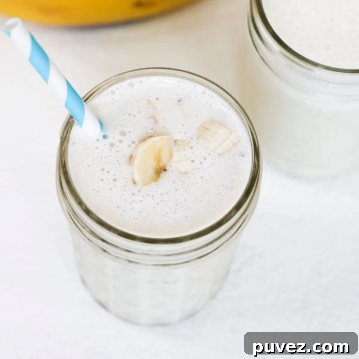 A creamy banana milkshake in a glass, topped with fresh banana slices and served with a blue and white striped straw. It looks inviting and ready to drink.