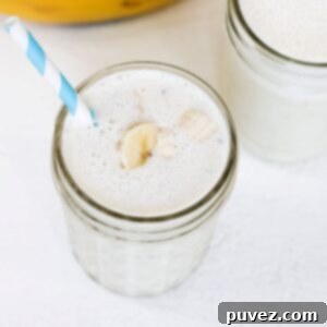 A tall glass filled with a creamy banana milkshake, topped with a few banana slices and served with a blue and white striped straw. It looks delicious and ready to be enjoyed.