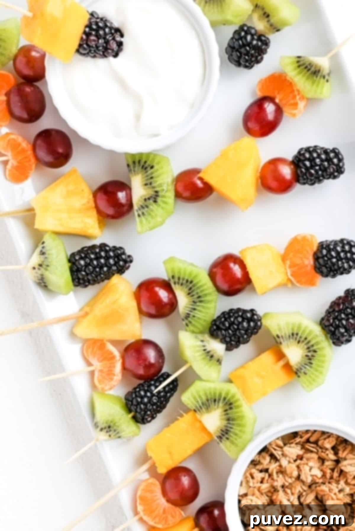 The Ultimate Fruit Skewers 2 Colorful pieces of fresh fruit on toothpicks and longer bamboo skewers, artfully arranged on a white plate, accompanied by small ramekins of granola and creamy yogurt for dipping. A perfect healthy snack or appetizer for kids and adults alike.