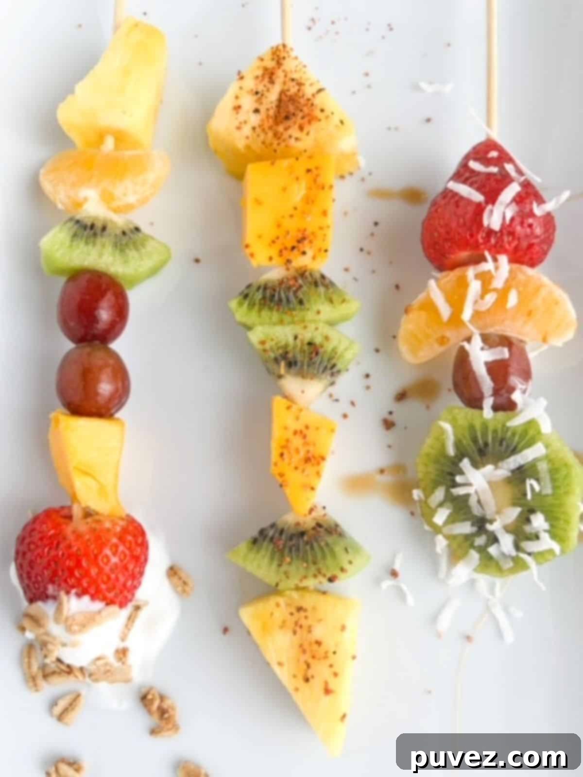The Ultimate Fruit Skewers 3 Three colorful fruit skewers displayed on a pristine white plate, each adorned with a distinct and appealing topping. One skewer features a drizzle of honey and a sprinkle of shredded coconut, another is coated in a vibrant dusting of Tajin seasoning, and the third is served alongside a small bowl of creamy yogurt and crunchy granola, showcasing diverse serving suggestions.