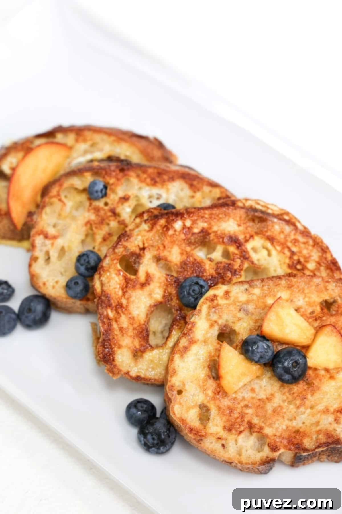Four golden-brown slices of sourdough French toast overlapping, beautifully garnished with fresh blueberries and juicy peach slices, ready for a delightful breakfast.