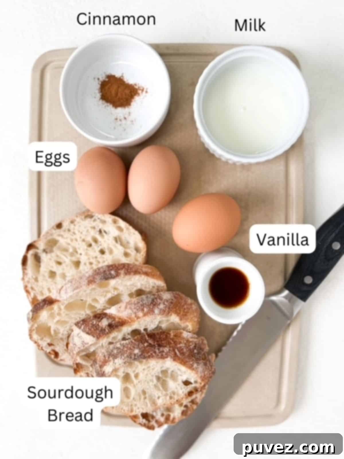 Individual ingredients for Sourdough French Toast arranged neatly on a cutting board: slices of sourdough bread, a glass of milk, eggs, a bottle of vanilla extract, cinnamon powder, and a bread knife.