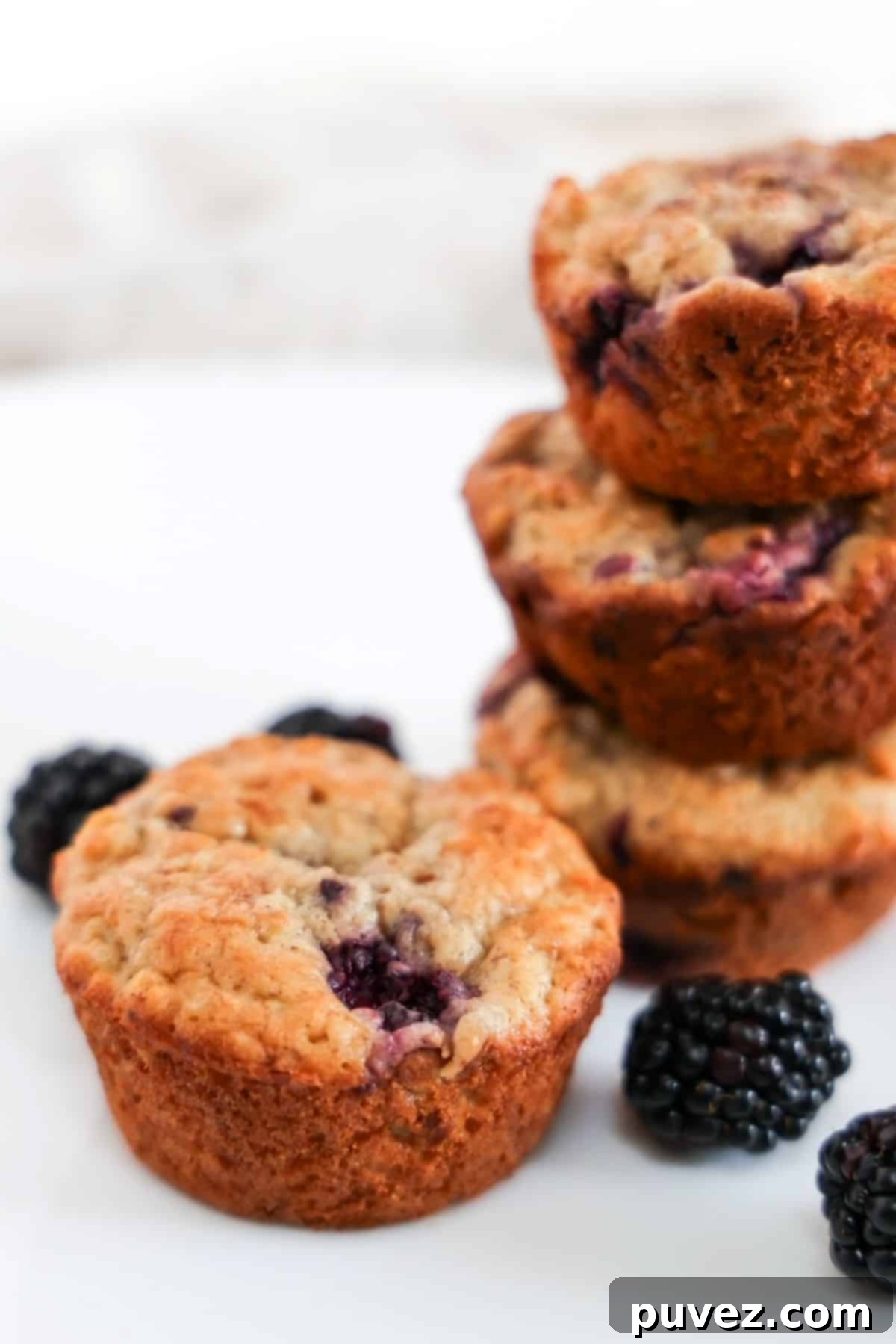 Four delicious banana blackberry oatmeal muffins. Three are artfully stacked to the right, with one perfectly placed in the center. Fresh, vibrant blackberries are scattered around, highlighting the key fruit ingredient.