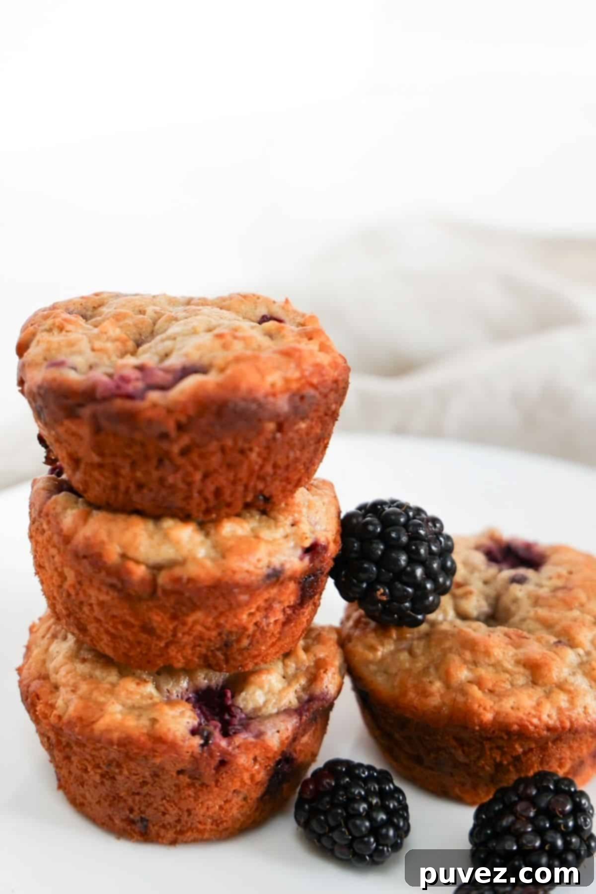 Four wholesome Banana Blackberry Oatmeal Muffins, artfully arranged with three stacked on the left and one individual muffin standing proud. A scattering of fresh blackberries adds a touch of natural beauty to the scene.