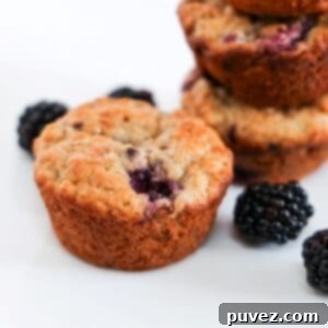 One delicious Banana Blackberry Oatmeal Muffin prominently in the foreground, with a scattering of fresh blackberries and a stack of other muffins blurred in the background, inviting a bite.