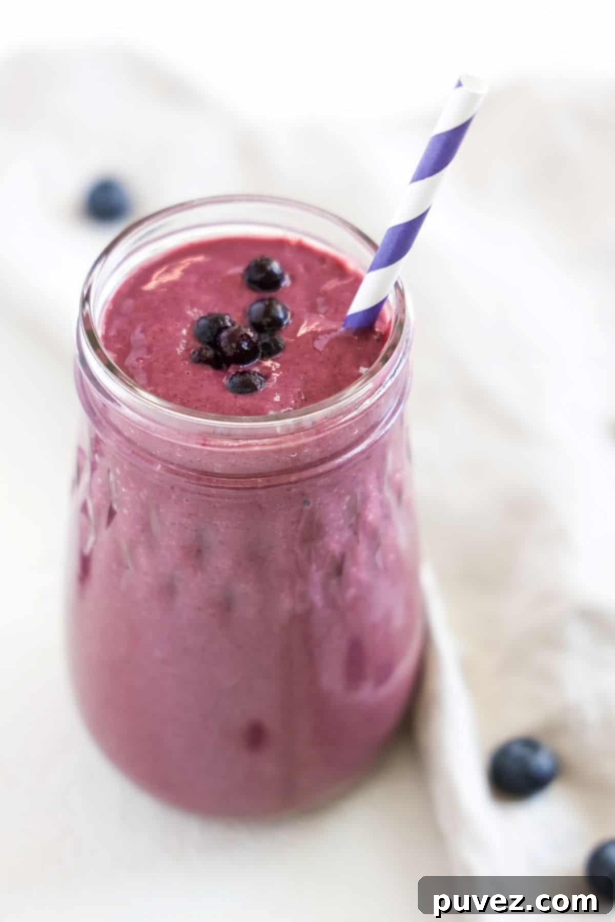 Blueberry tofu smoothie served in a glass jar with a purple and white striped straw surrounded by a linen napkin and a few blueberries.