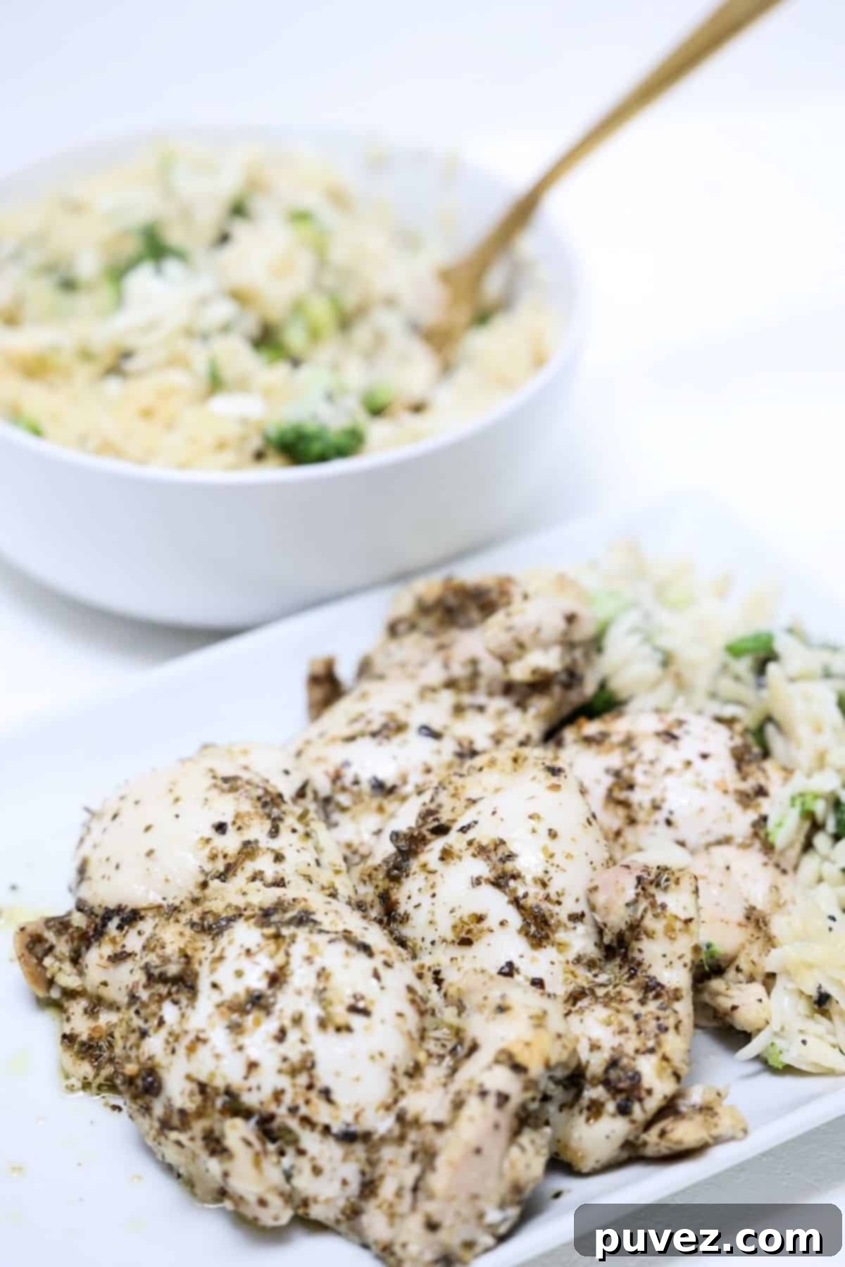Pieces of baked Italian chicken thighs on a white plate with a bowl of orzo in a white bowl behind the plate, garnished with fresh herbs.