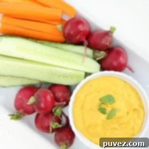 Carrot hummus in a white ramekin with raw veggies on a white rectangular plate.