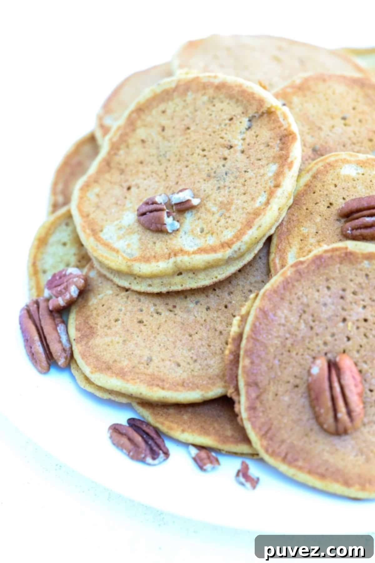 A pile of pumpkin pancakes on a white plate with pecans scattered around them.